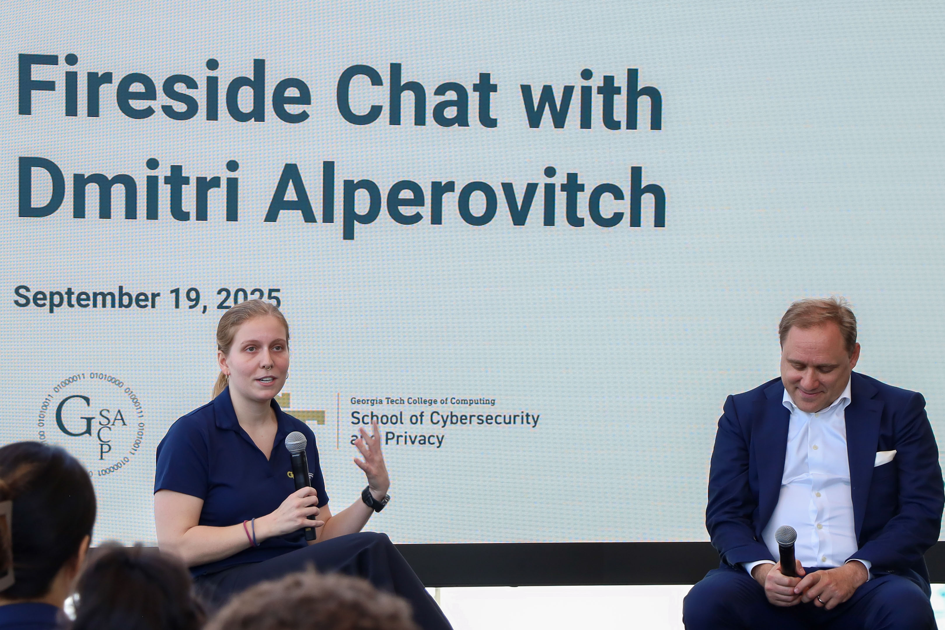A blonde woman in a blue polo speaks into a microphone and gestures with her hand. Next to her is a man with blonde hair wearing a blue suit and holding a microphone. Behind them is a large screen that says, "Fireside Chat with Dmitri Alperovitch. September 19, 2025." The School of Cybersecurity and Privacy logo can be seen behind the woman and the SCP Graduate Student Association next to her shoulder.