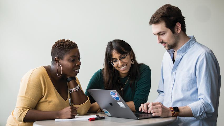 Three students, two women and one man, stand huddled by a lap top.