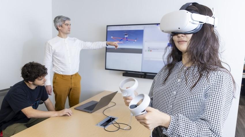 A female student wears a Meta Quest VR headset with two men behind her